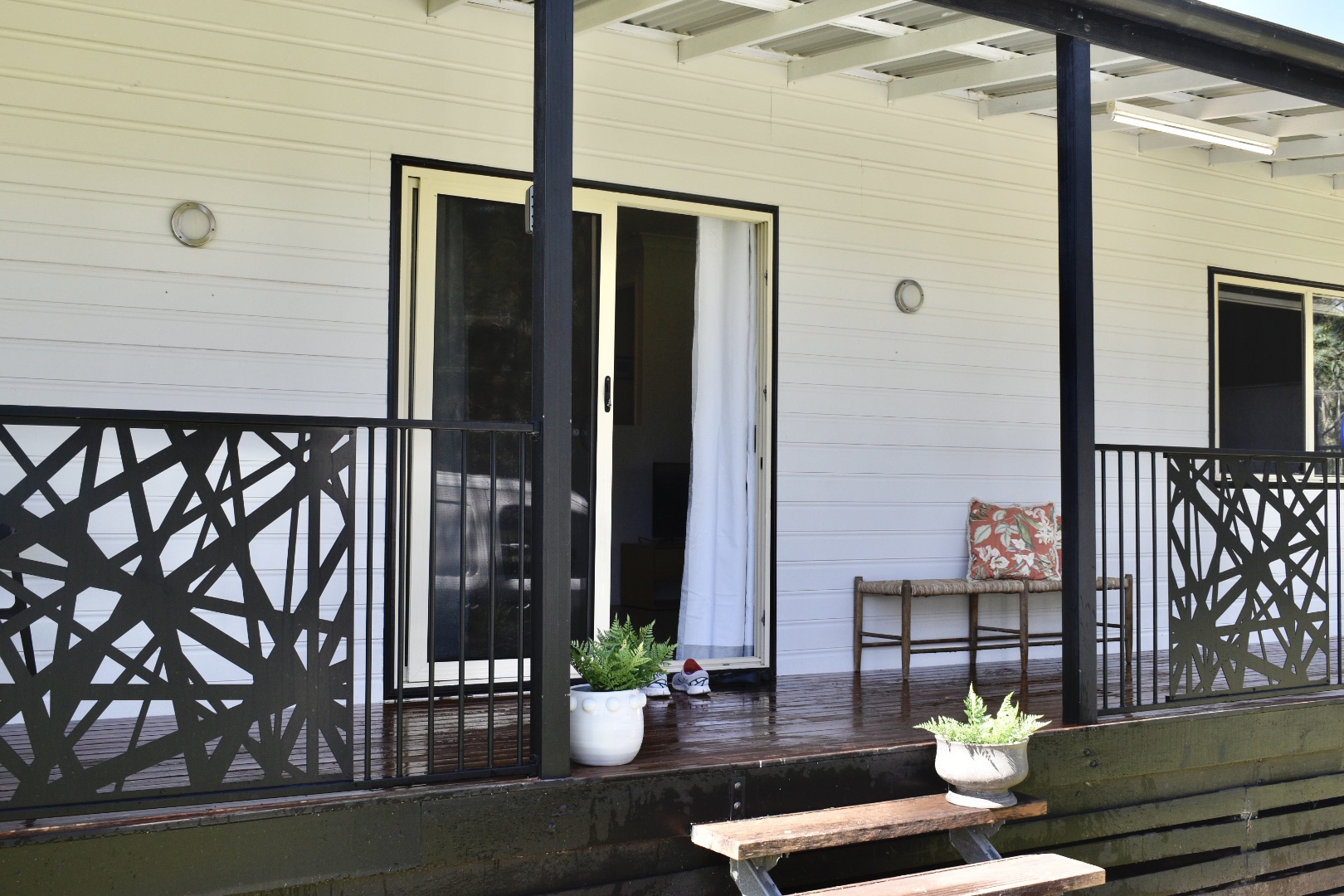 A home's entrance, including stairs up to a wooden deck, and a sliding door. There are pot plants on the deck.