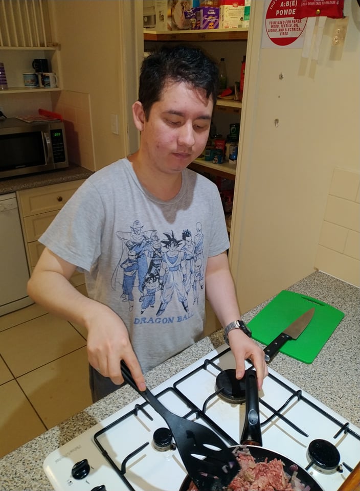 Ronan stands at the kitchen stove with a frypan of mince in front of him. He is carefully holding the pan's handle in one hand, and using a spatula to stir the mince.