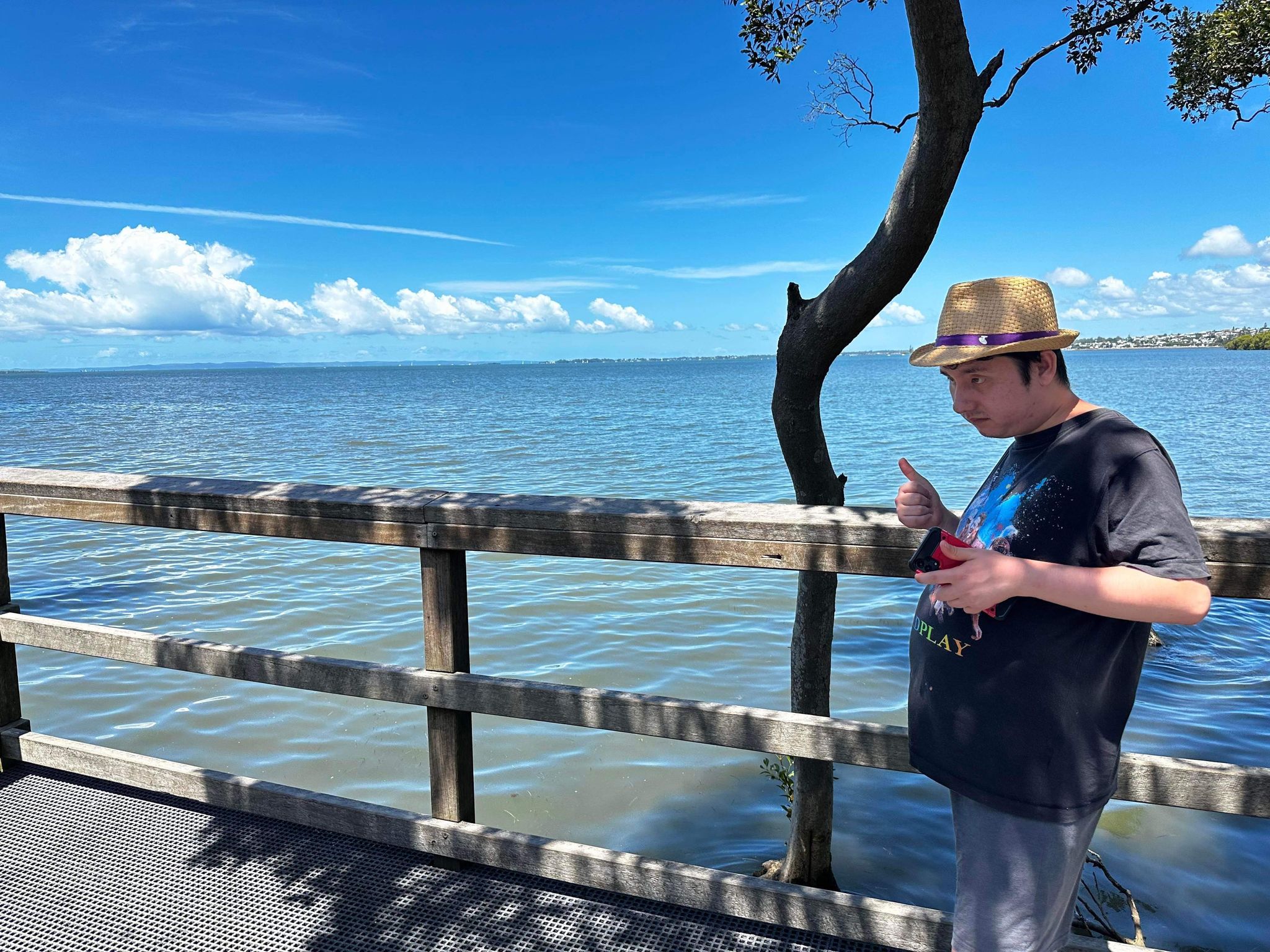 A man stands on a jetty with the ocean behind him and a bright blue sky. He is giving the thumbs-up sign.