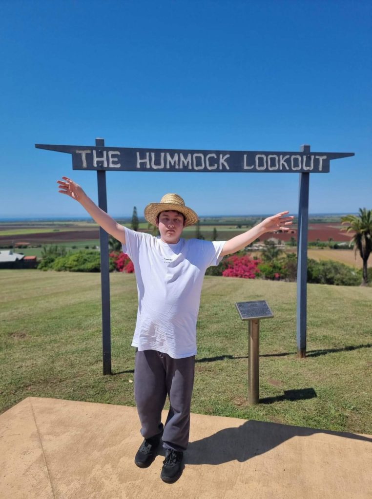 Ronan, a young man, stands with arms in the air in front of a sign saying "The Hummock Lookout". There are gardens behind him and country farmland in the background.