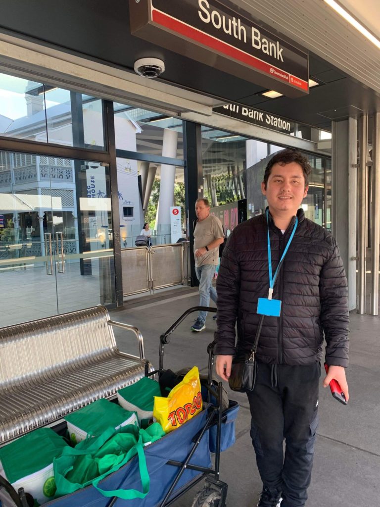 Ronan stands happily at South Bank train station. He has a wagon packed with bags next to him.