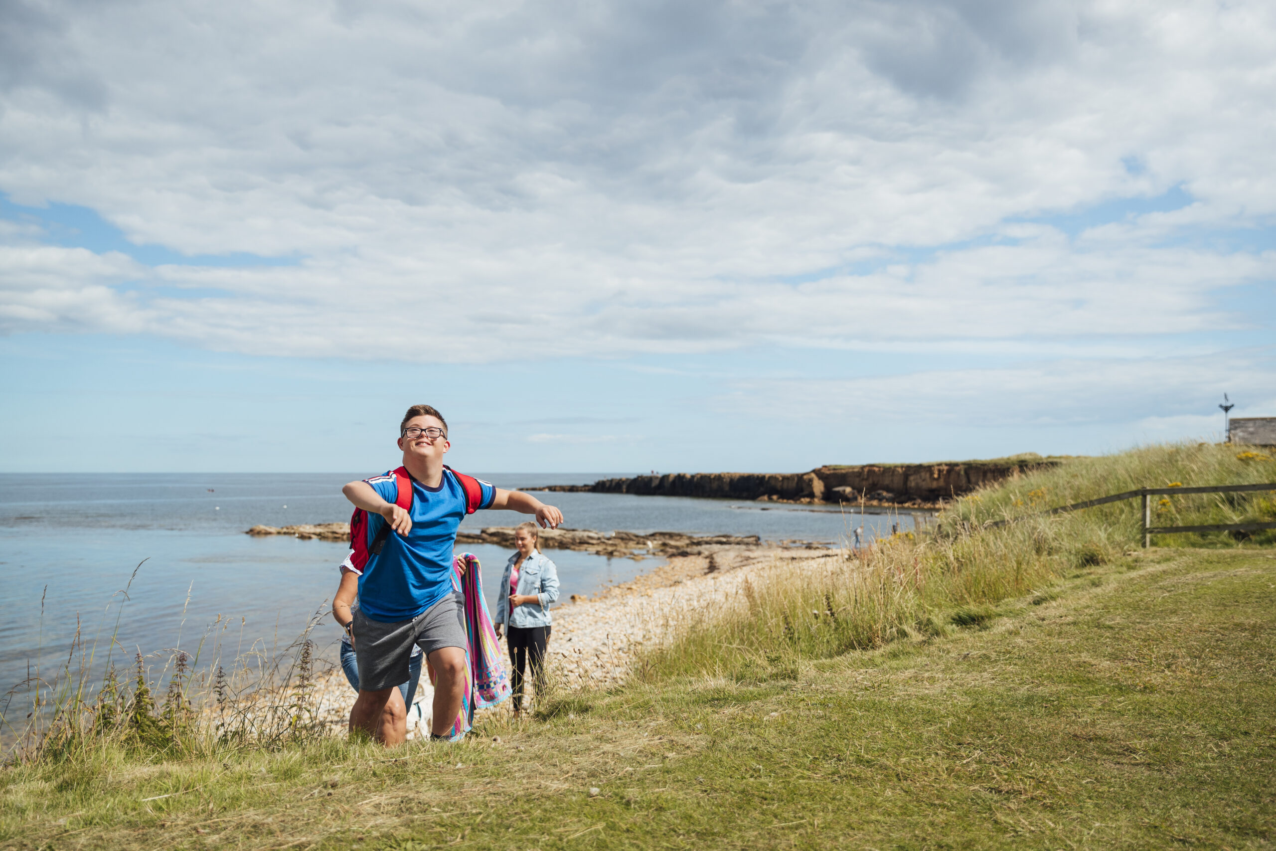 A view of a coastline with sand dunes in the foreground and the ocean in the background. A young man with Downs Syndrome has hiked up the sand dunes. He looks energetic and triumphant. Two other people are hiking up behind him.