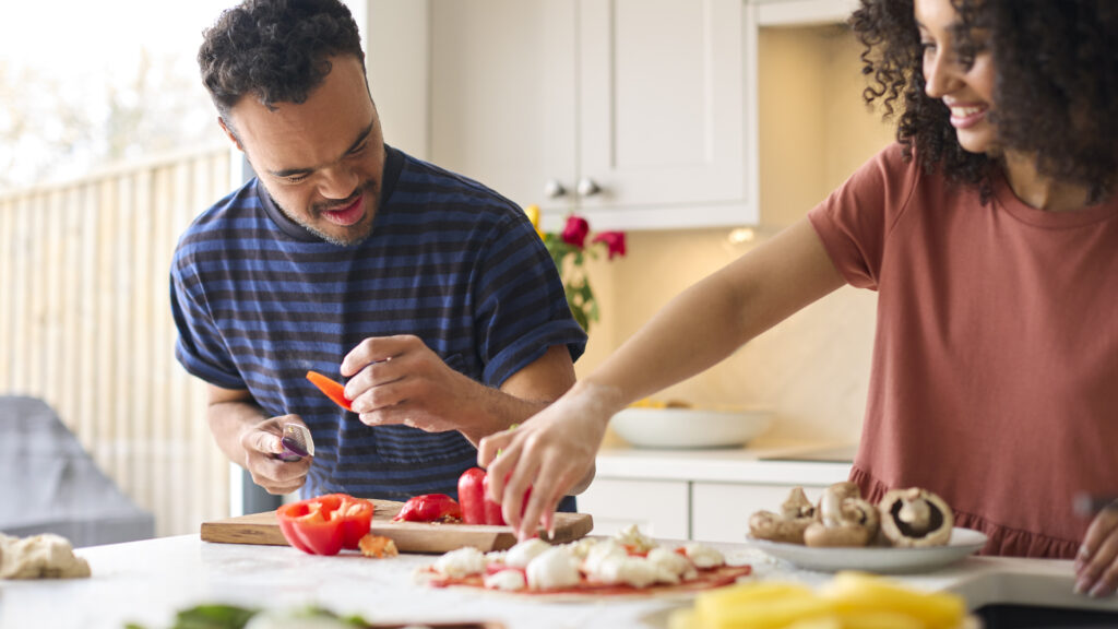 A man with disability is in a kitchen chopping capsicum and making a pizza. A woman is helping him.