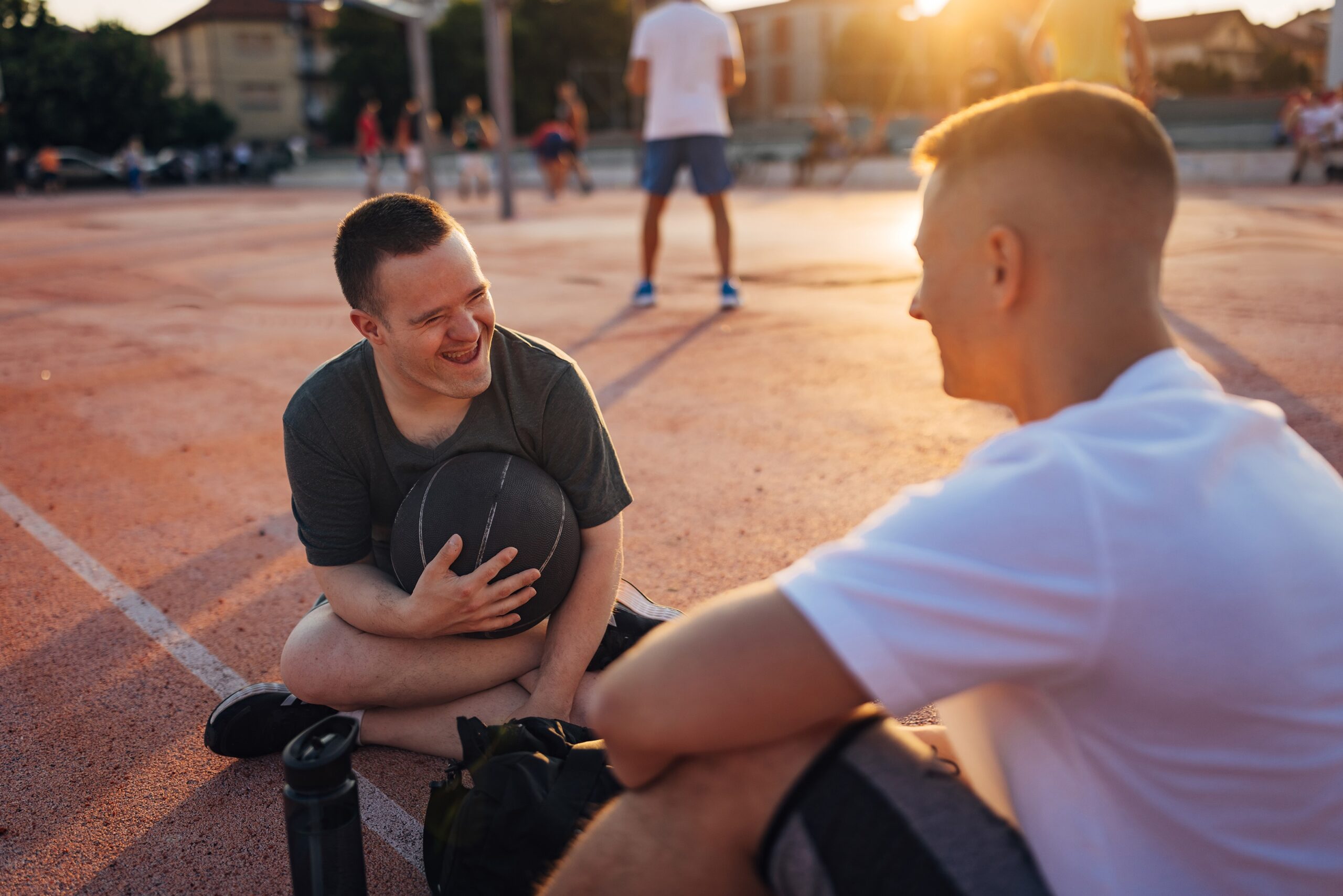 Two men sit on a basketball court in the late afternoon chatting and laughing. One of the men, who has Downs Syndrome, holds a basketball in his lap.