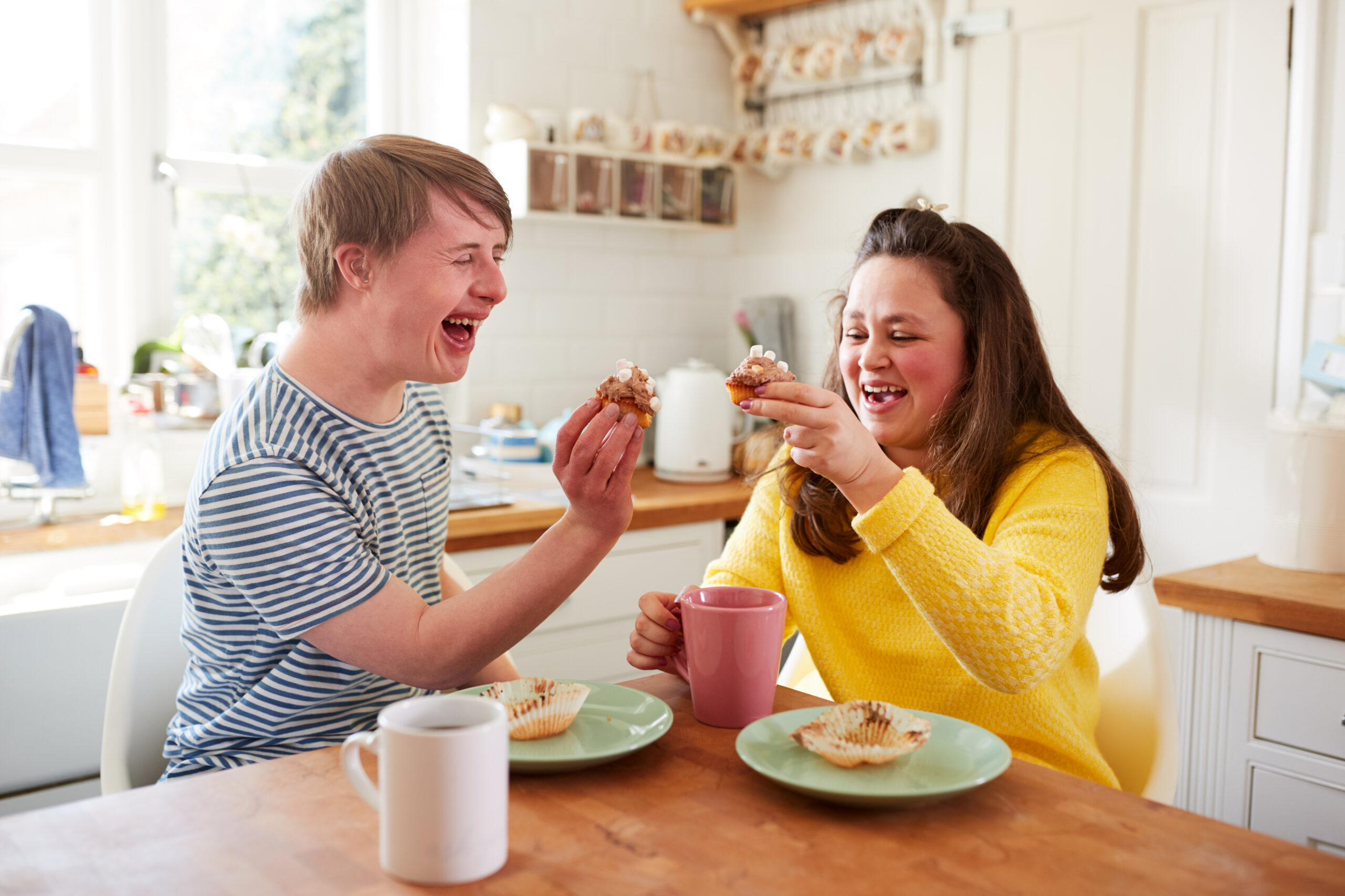 A young man and woman with Downs Syndrome sit together at a kitchen table, enjoying tea and muffins.